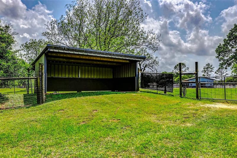 Open-front metal loafing shed on a green rise in a fenced pasture, with a metal gate and trees behind it