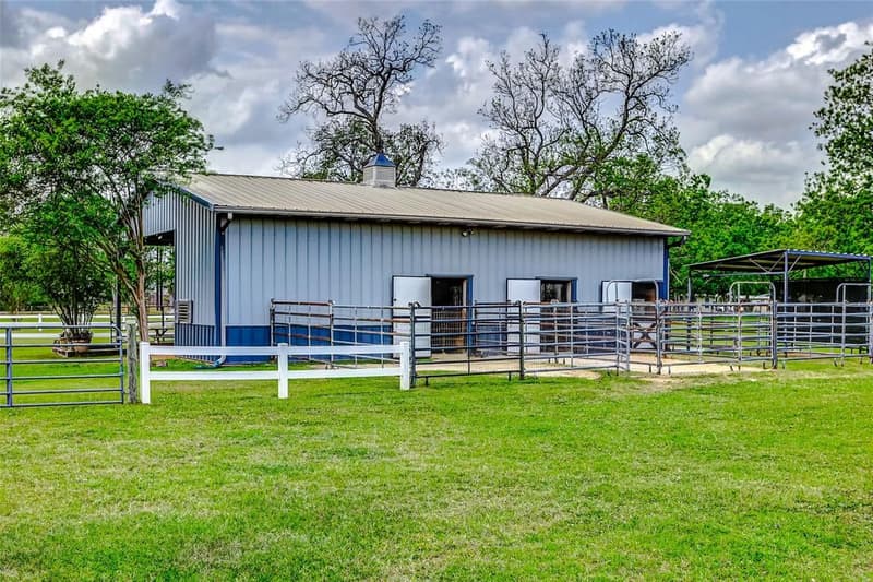 Gray metal horse barn with blue trim and a cupola, open stall doors and pipe-panel paddock runs along its side, pasture in front