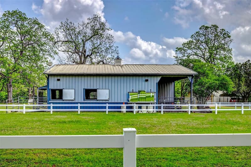 Side view of a gray and blue metal horse barn with stall windows, cupola and covered area, seen across white rail fencing