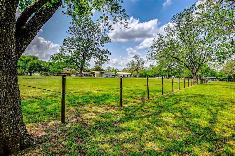 Broad green pasture with a wire fence line and tall shade trees, barns, carports and the brick home in the distance