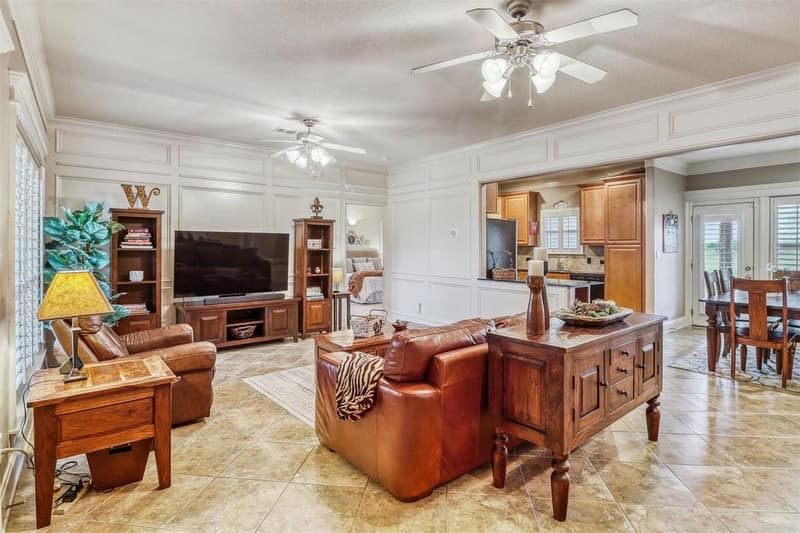 Living room with wainscot paneled walls, crown molding, two ceiling fans and plantation shutters, open to the kitchen