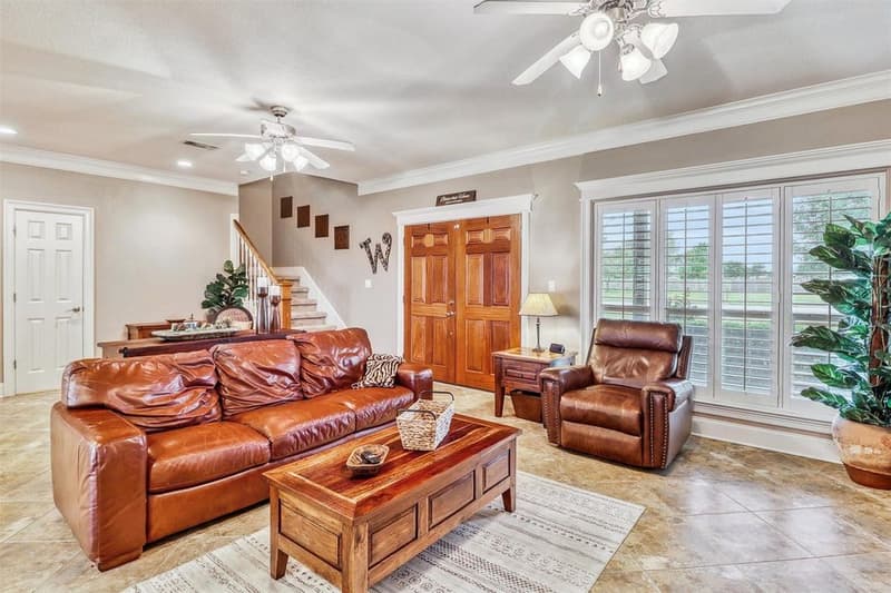 Living room with tile floors, wood double front doors, staircase and a wide window with plantation shutters