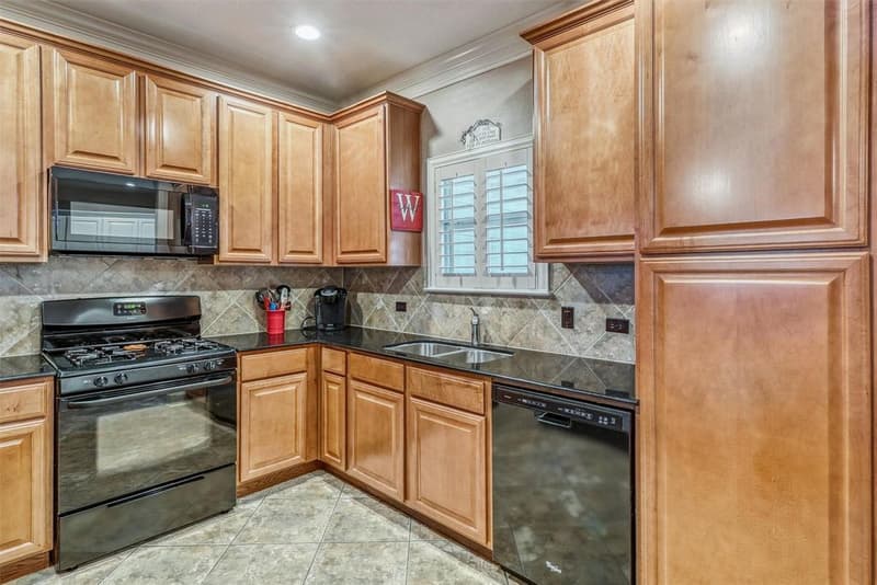 Kitchen corner with wood cabinets, dark granite counters, double sink under a shuttered window, gas range, dishwasher