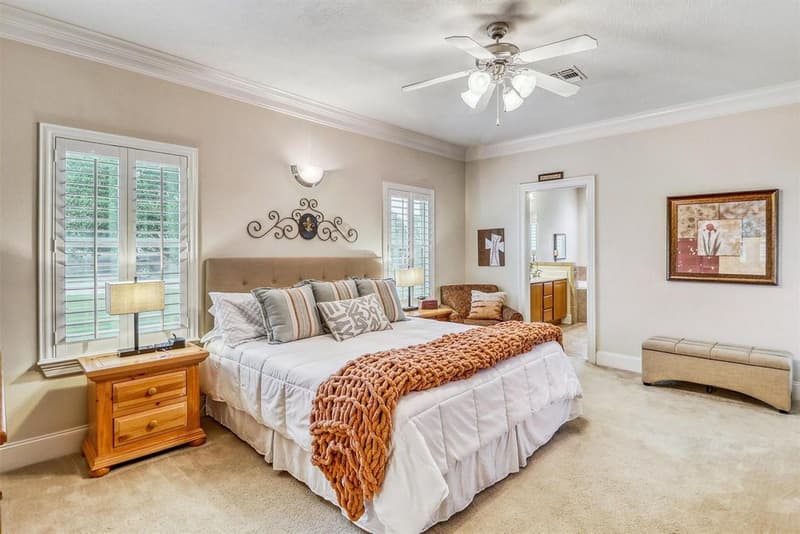 Primary bedroom with carpet, crown molding, ceiling fan, plantation shutters and a doorway to the primary bath