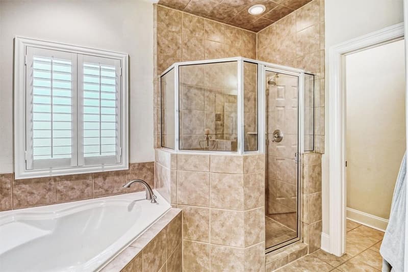 Tiled corner shower with glass enclosure beside a soaking tub under a shuttered window