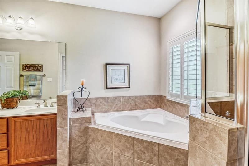 Corner soaking tub with tile surround and plantation shutters, vanity and mirror to the left