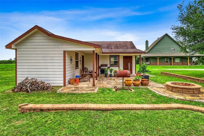 Side view of the detached building with porch, flagstone patio, fire pit and stacked firewood on an open lawn