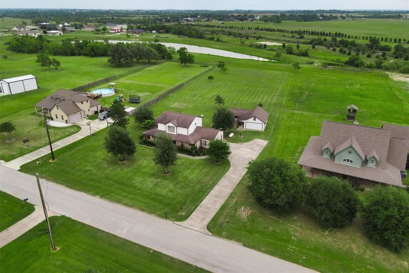 Drone view from the street of the home, long driveway and detached garage building, with open fields and water in the distance