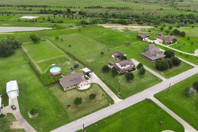 Wide drone view of nearby acreage homesites along the street, open pasture and a pond in the distance