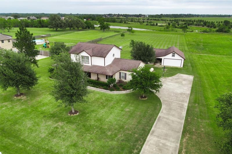 Low drone view of the two-story home's front, mature trees, curving driveway and detached garage building, with a pond on the horizon