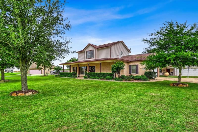 Front of the two-story brick and siding home with a covered front porch, trimmed hedges and a wide green lawn under blue sky